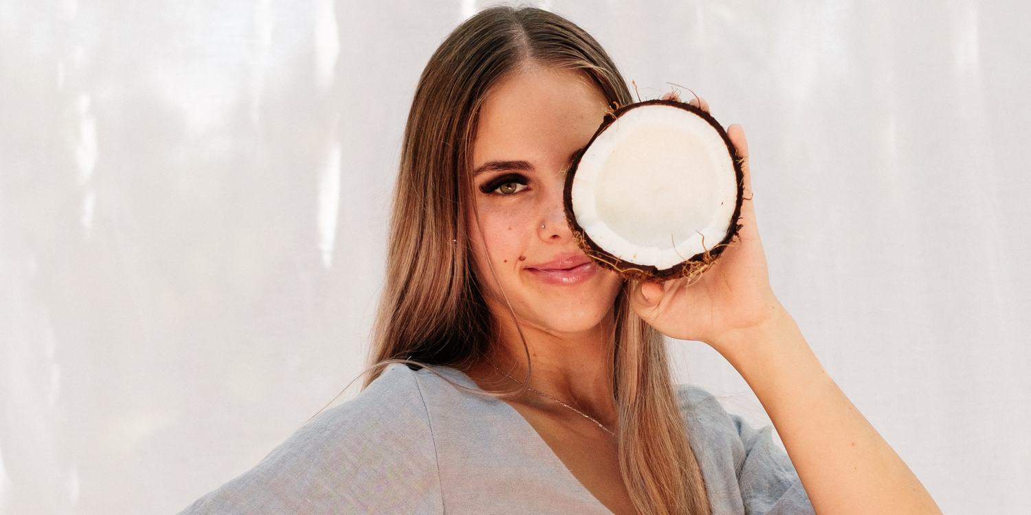 Pretty young long brown hair girl holding half a freshly cut coconut up in front of her left eye while smiling lips closed, brown eyes opened. wearing a grey linen bell sleeve shirt.  Standing in front of a white linen sheet in the sun shine with shadows.